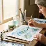 Person malt mit Aquarellfarben abstrakte Formen in Blau und Grün auf Papier, Pinsel und Farben im warmen Licht verstreut